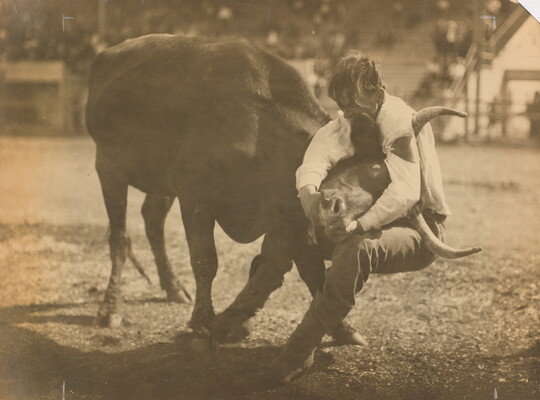 A sepia-toned photograph of a man with his arms wrapped around a bull's head from behind.