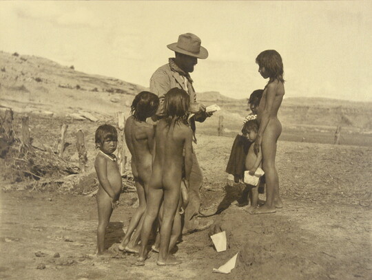 A sepia photograph of a group of mostly unclothed Indigenous children gathered around a White man in a barren landscape.