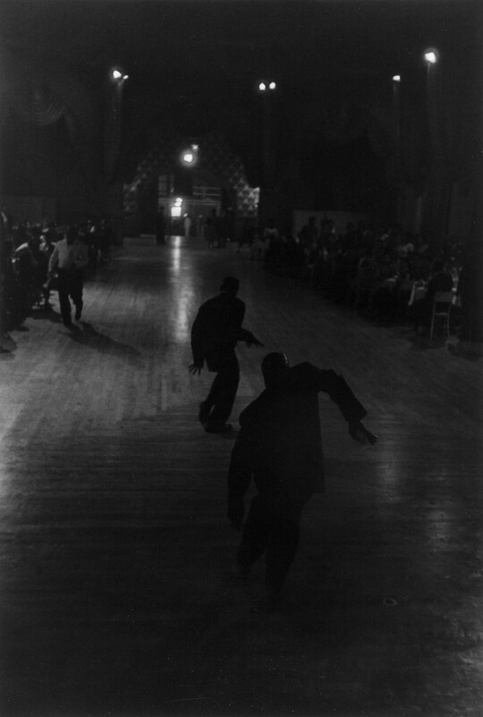 A dark black-and-white photograph of two male dancers in silhouette at the center of a dance floor.