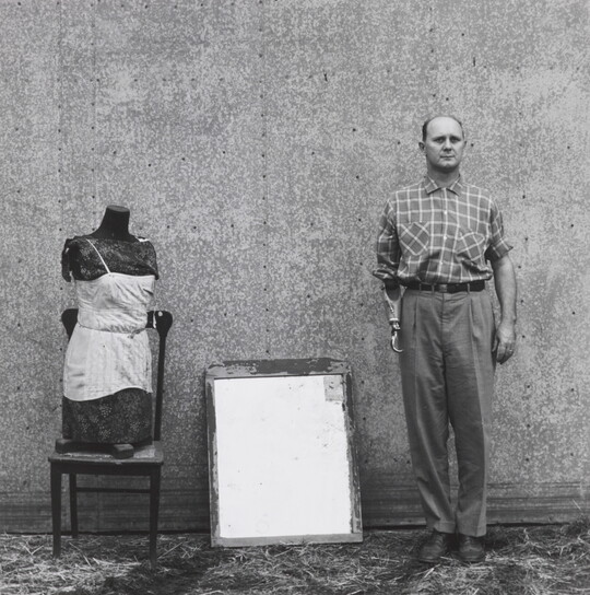 A black-and-white photograph of a mannequin bust on a wood chair, a mirror, and a balding White man with a prosthetic arm that ends in a hook lined up against a wall.