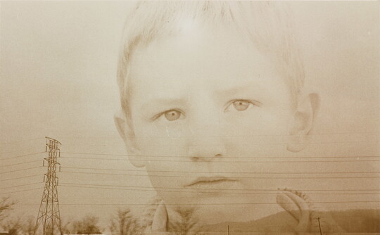A sepia-toned double-exposed photograph of the face of a young White child over a landscape of powerlines and hills.