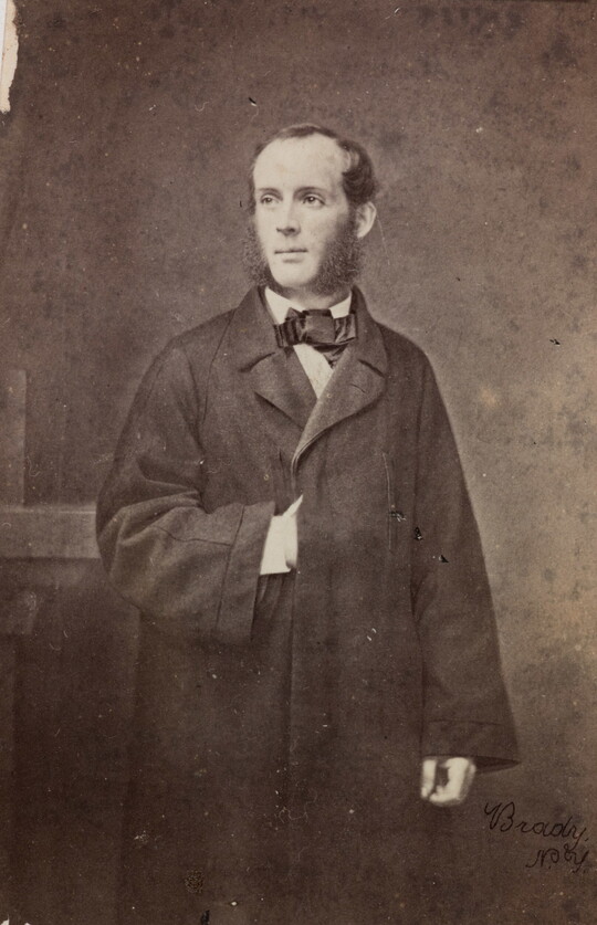 A black-and-white photograph of a White man with large sideburns wearing a long coat; one hand tucked between the buttons.