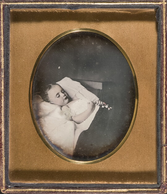 A black-and-white photograph in a gold frame of a child with closed eyes laying in bed with a toy in her clasped hands.