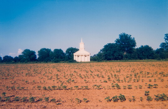 A color photograph of a white wood church with a tall steeple and columns on the front sitting across an open field.