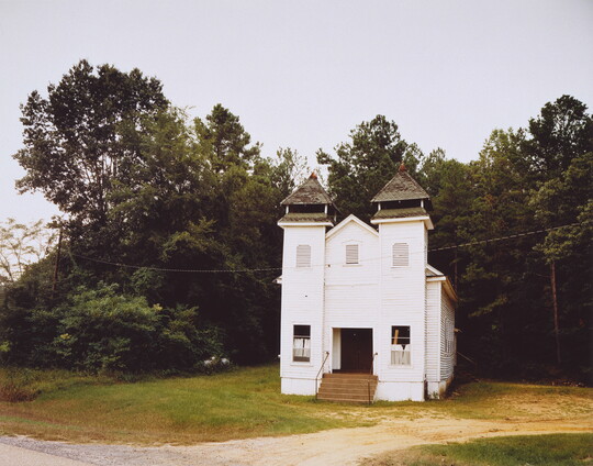 A color photograph of a white chapel situated in front of a wood.