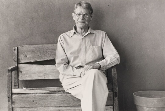 A black-and-white photograph of an older White man wearing glasses and a button-down shirt sitting on a wood bench.