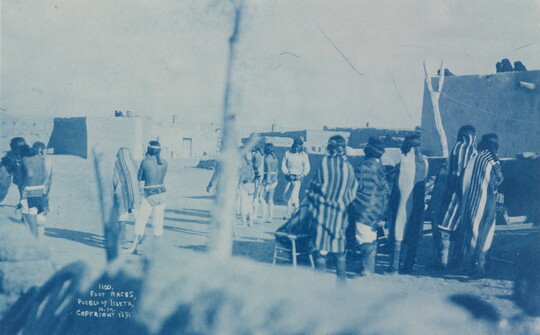 A blue-tinted photograph of Native American people, some wrapped in blankets, on a dirt road; several adobe buildings nearby.