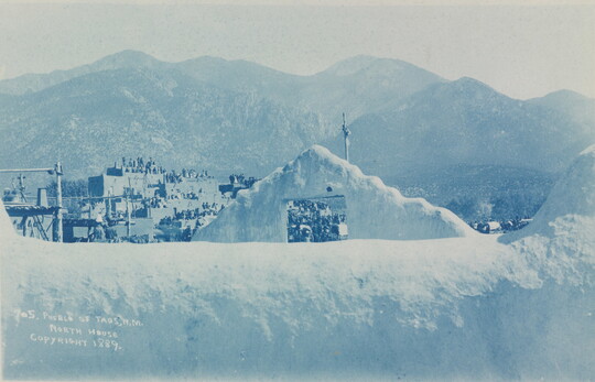 A blue-tinted photograph of a multi-story pueblo at the base of mountains as seen from over an adobe wall.