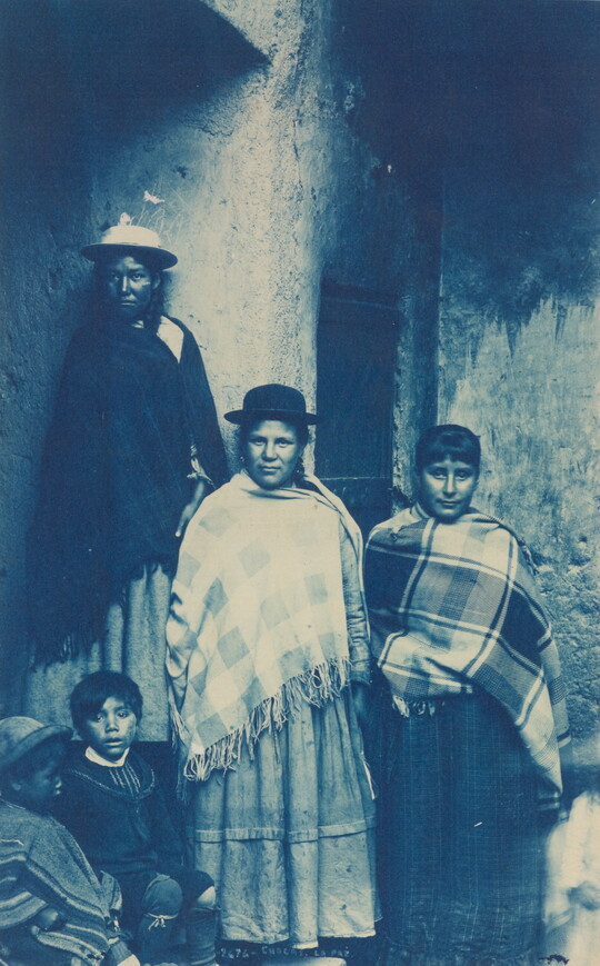 A blue-tinted photograph of women and children wearing ponchos standing next to a wall.
