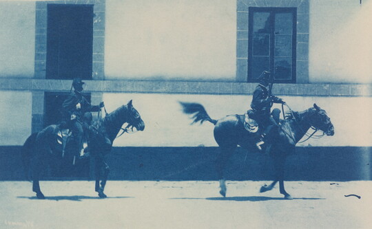 A blue-tinted photograph of two men in uniforms riding horses past a building.