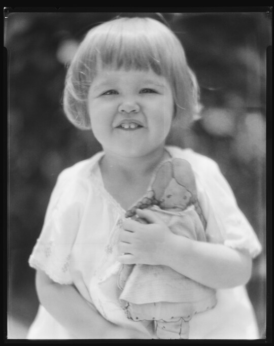 A black-and-white image of a young White child with short hair, showing her two front teeth, and holding a doll.