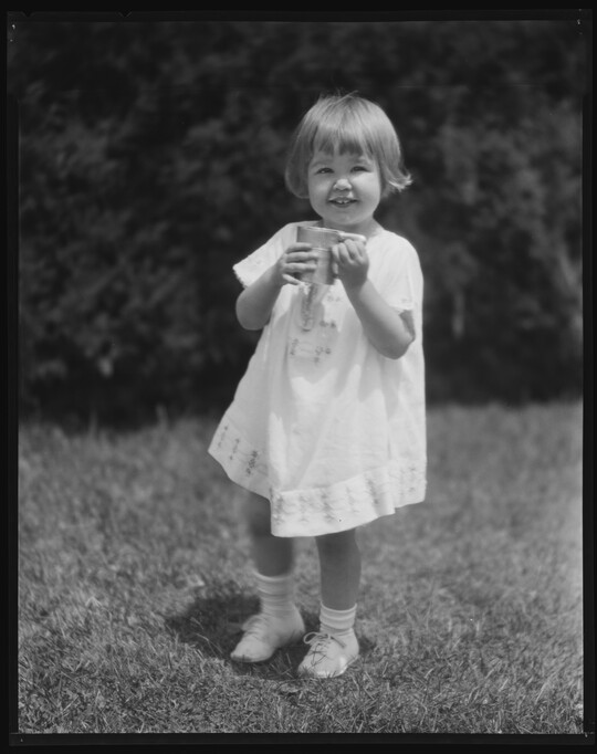 A black-and-white image of a young White child with short hair wearing a white dress and holding a mug as she stands in the grass.