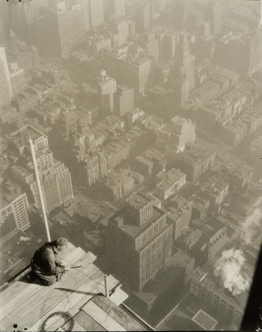 A black-and-white photograph taken from above of a man kneeling precariously on the edge of a construction platform suspended high over a city skyline.