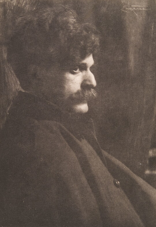 A black-and-white photograph of a White man with lots of dark, curly hair and a mustache sitting in profile.