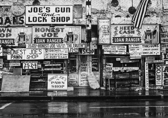 A black-and-white photograph of the outside of a pawn shop covered in signs advertising "Honest Joe" and the "Loan Ranger."