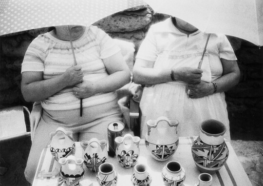 A black-and-white photograph of two women, faces hidden by umbrellas they are holding, at a table filled with pottery decorated with Native American designs.