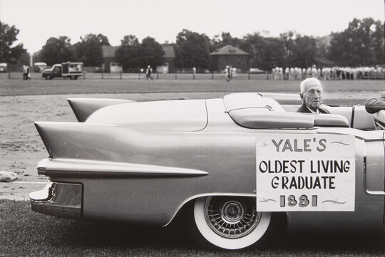A black-and-white photograph of an elderly White man seated in the back of a convertible; a sign on the car reads, "Yale's oldest living graduate, 1881."