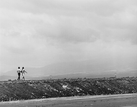 A black-and-white photograph of two figures walking along high berm with water below.