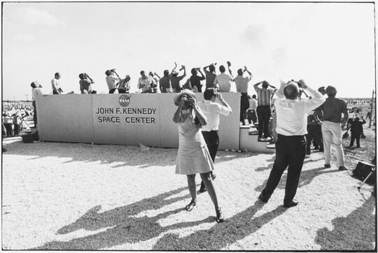 A black-and-white photograph of crowd of people looking up into the sky from a viewing stand on a beach with a sign that reads, "John F. Kennedy Space Center."