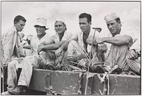 A black-and-white photograph of five young White men in dirty work clothes sitting in a wagon.