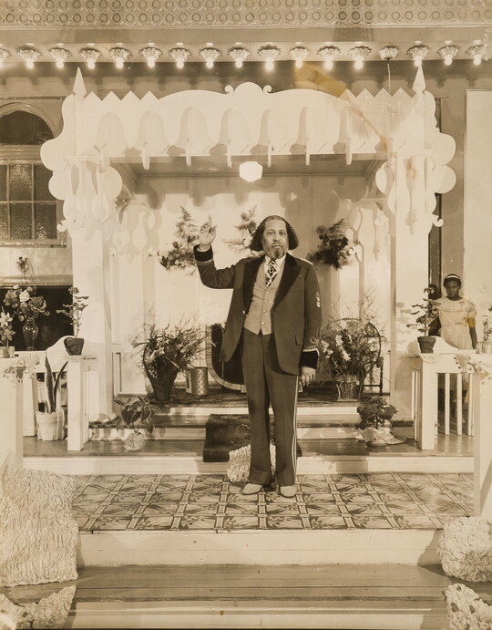 A sepia-toned portrait photograph of a Black man with mid-length coiled hair, in a three-piece suit with his arm raised and index finger pointing up, and an altar with flower arrangements behind him.