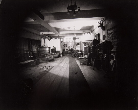 A dark black-and-white photograph of a living room with a spotlight on a man seated in an armchair in a far corner.