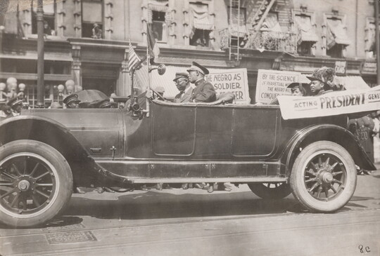 A black-and-white photograph of a convertible vehicle on an urban street with four Black people in the car, a banner draped on the side, and signs displayed in the background.