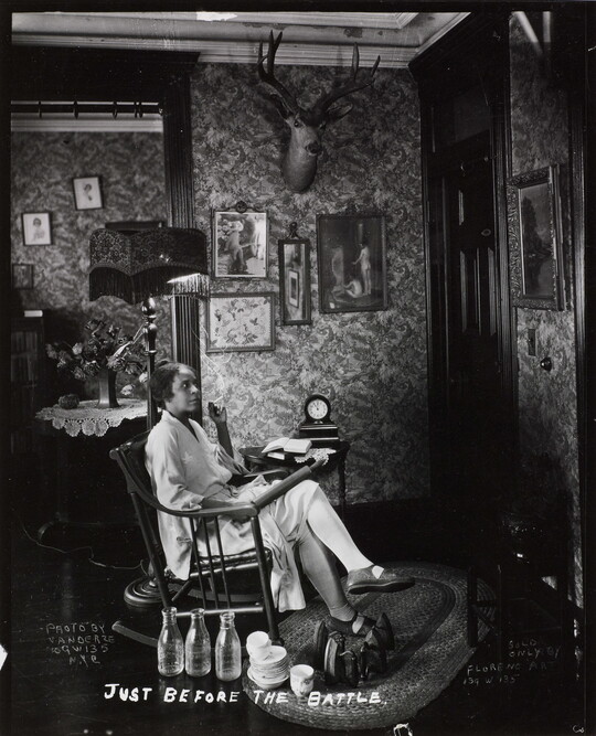A black-and-white photograph of a Black woman in a rocking chair, holding a rolling pin on her lap, waiting at a front door.