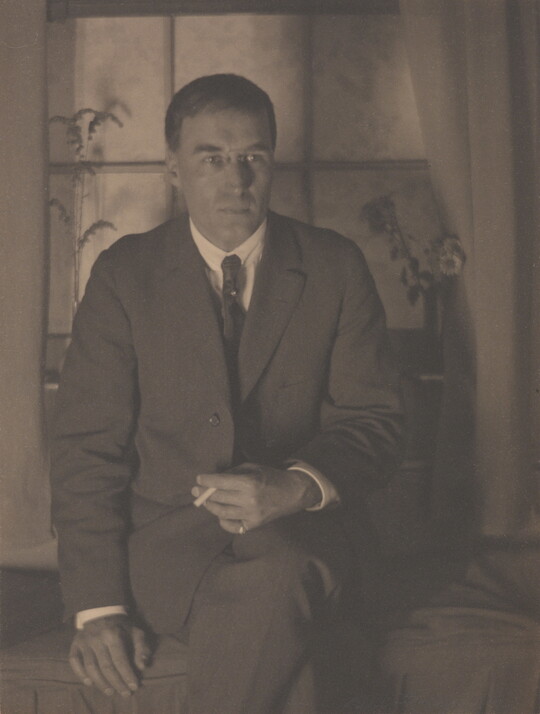 A sepia-toned photograph of a White man in a suit, legs crossed, holding a cigarette in one hand that is resting on his leg, seated in front of a window.