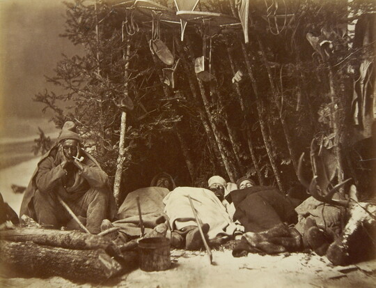 A black-and-white photograph of several men asleep outside at a winter campsite, with their snow gear stored in branches of trees.
