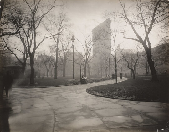 A black-and-white photograph of a park with bare trees and a tall building rising up in the distance.