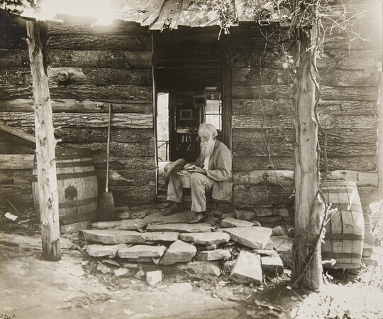 A black-and-white photograph of an old White man with a long beard sitting in the doorway of a log cabin.