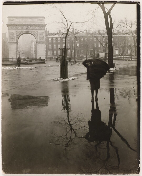 A black-and-white photograph of someone walking on wet pavement through a plaza with bare trees and a white arch in the distance.