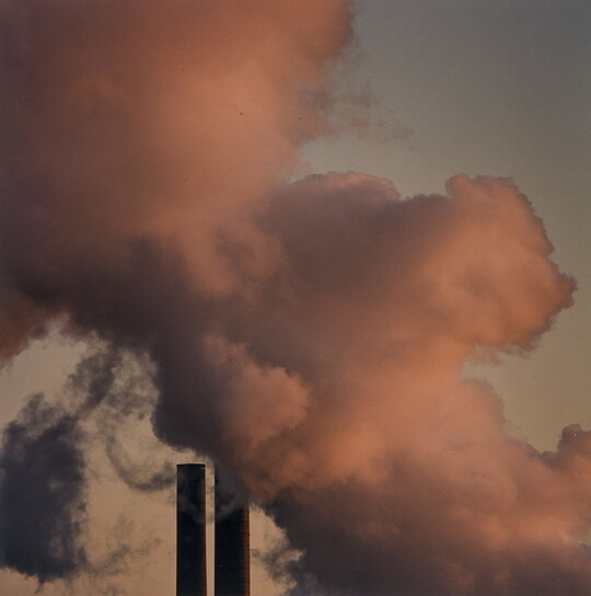 A color photograph of pink smoke against a gray sky above two industrial smokestacks.
