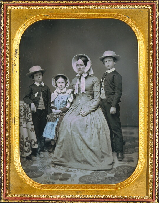 A hand-painted black-and-white formal portrait photograph in a gold frame of a White woman sitting with two boys and a girl standing around her.