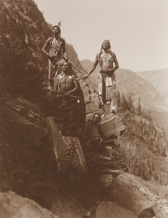 A sepia-toned photograph of four Native Americans in traditional dress arranged on a steep, rocky slope.