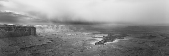 A black-and-white panoramic photograph of a steep canyon under a stormy sky.