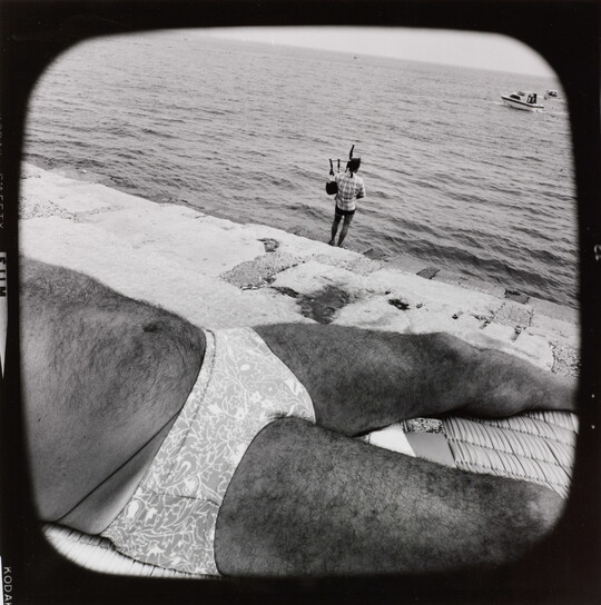 A black-and-white photograph of the hairy torso of a man wearing a brief-style swimsuit sunbathing near a cement ledge by water where a fully-clothed person plays bagpipes in the background.
