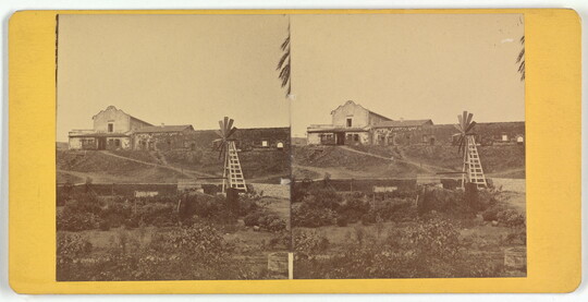Two almost identical side-by-side sepia-toned photographs of an adobe building on a hill with a windmill in the foreground.