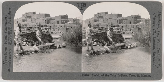 Two almost identical side-by-side black-and-white photographs of two Native Americans standing in front of a pile of mud bricks with an adobe pueblo in the background.