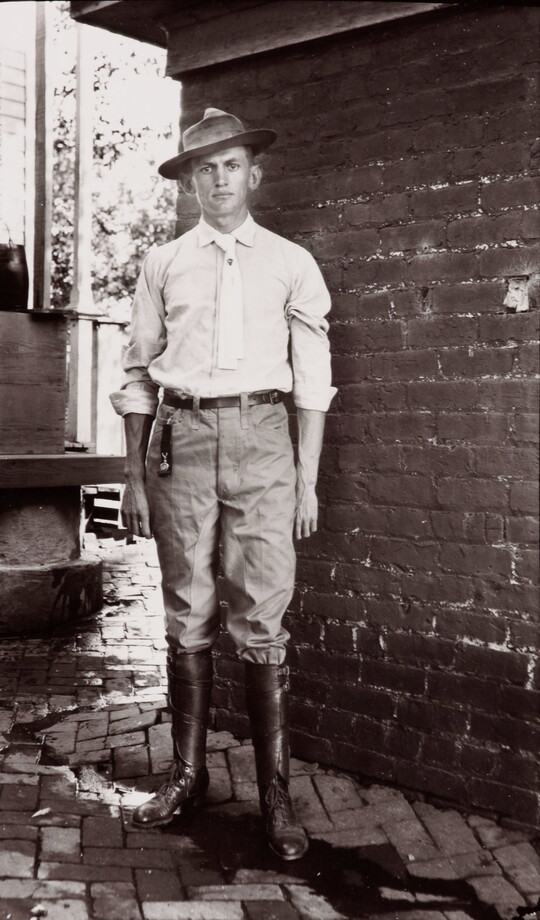 A black-and-white photograph of a young White man in a brimmed hat, light shirt and tie, pants, and tall boots standing in front of brick wall.