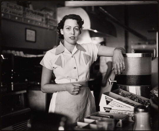A black-and-white photograph of a White woman in a waitress uniform standing behind a diner counter with an arm propped on drink dispenser.