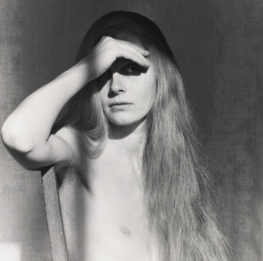 A black-and-white portrait photograph of a nude White woman with long hair using her hand to shield her eyes.