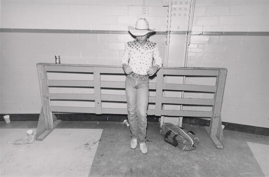 A black-and-white photograph of a teenager wearing a cowboy hat and a decorative western-style shirt leaning back against a showjumping fence.