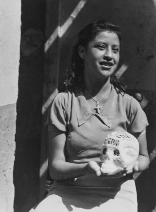 A black-and-white photograph of a brown-skinned girl showing the decorated sugar skull she holds in her hands.