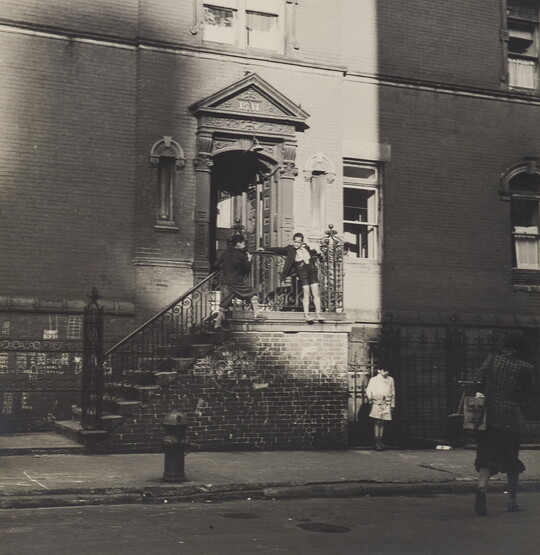 A black-and-white photograph of the entrance to an apartment building with children playing on the outside of the railing on the steps that lead to the door.