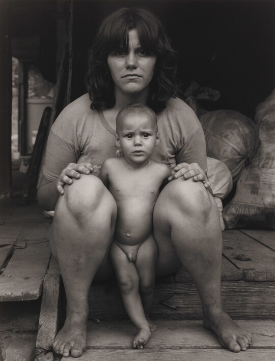 A black-and-white photograph of an expressionless White woman sitting on a wood step with a naked baby standing between her legs.