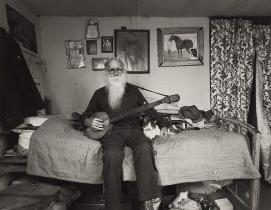 A black-and-white photograph of an elderly White man with a long white beard sitting on a bed in a cozy bedroom holding a banjo with one hand, the other hand around a cat lying next to him.