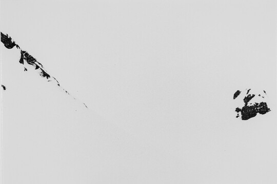 A black-and-white photograph of mountains totally covered with snow except for dark rocky formations in the upper left and center right.