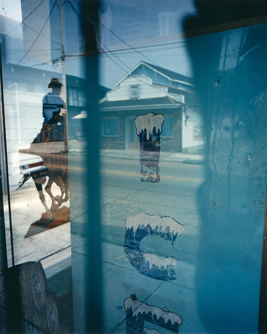 A color photograph taken through a window by the side of an ice machine and a poster of a man on a horse, as well as reflections of a house and other buildings.